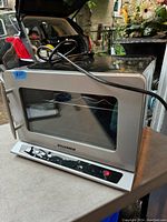Front and side view of the silver and black Sylvania wine fridge cooler with control buttons and power light visible on the base panel.