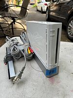 White Nintendo Wii console with power cable, AV cable, and sensor bar placed on outdoor table.