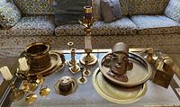 Various views of brass decor items displayed on a glass-topped table with brass frame including candlesticks, trays, decorative mask, bird figurine, and container.