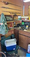 Fishing rods hung on a pegboard wall above wooden shelves with fishing related gear including tackle boxes, vest, net, and various small items. Cooler and camping chair visible below shelf.