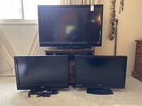 Three flat screen televisions arranged on floor and on a stand. Vizio TV is largest and placed on stand. Sony and Sylvania TVs on floor in front.