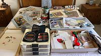 Full view of sewing and needlepoint items laid out on a bed, showing numerous embroidery canvases, organizers with threads, and small kits.