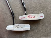 Close-up of two white Coors branded putters showing logos and head shapes