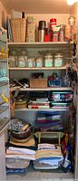 Wide view of pantry closet shelves containing jars, linens, cookbooks, thermoses, tins, baskets, and miscellaneous kitchen items