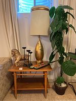 Side view of mid century wooden side table, gold 1950s lamp, black candleholders, wooden horse figurine, and decorative wooden wall piece arranged on and around the table beside a sofa and plastic leafy plant in a basket.