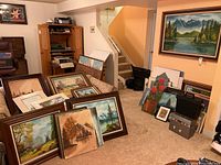 Overview of living room area with numerous framed paintings leaning against couches and walls, showcasing landscape and nature themes
