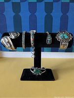 Overall display of six items on black T-bar stand: plain cuff, turquoise bead cuff, watch band, triangular inlay cuff, large turquoise cabochon cuff, separate malachite cuff