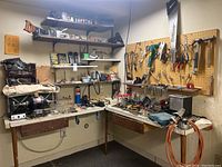 Wide-angle photo showing corner workshop with workbenches, pegboards holding various hand tools, tool boxes, power tools, and supplies on shelves.