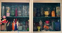 Photo shows cabinet filled with assorted glass and ceramic vases, pitchers, and decorative containers in various colors. Also visible are small bowls, candle holders, and bottles in different shapes and sizes.