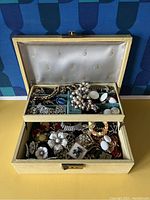Open jewelry box showing lower drawer filled with mixed vintage costume jewelry pieces such as floral brooches and chain bracelets in various tones.