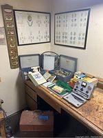 Photo showing two blue dental equipment cases opened on a wooden workbench with various dental instruments inside, a gray metal case with dental tools and dials, manuals and books, and a small green pouch. Above the bench are framed dental anatomy diagrams and a vertical wooden sign reading 'DENTURES' with an image of teeth.