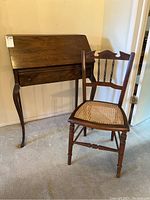 Photo showing the vintage wooden drop leaf desk with a single drawer and the wooden cane seat chair beside it, both standing on carpeted floor.