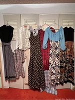View of 7 vintage clothing items hanging on hangers including dresses, tops, and a vest with various patterns and colors.