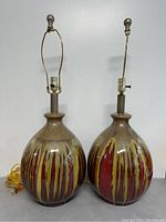 Front view of both lamps showing full height including harp and finial, and ceramic bases with yellow and red vertical drip stripe glaze over mottled brown ceramic.