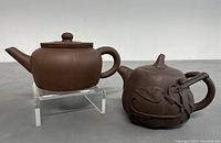 Front side view of both teapots on a gray surface, showing the different designs: one smooth, one textured with leaves and vines.