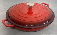 Closed red enameled cast iron pan with lid and double side handles, showing silver knob and glossy finish