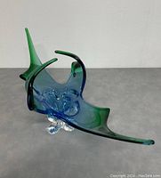 Side angle view of free form art glass bowl with green to blue color gradient and clear base on gray surface.