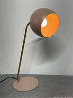 Front view of the lamp lit showing the round pink base, brass arm, and globe shade with orange interior lighting the bulb.