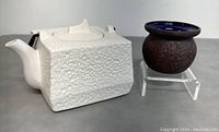 White textured MCM teapot with black handle and small brown bowl/candle holder side by side on gray surface.