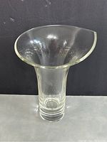 Front view of clear glass vase showing wide flared asymmetrical rim and thick cylindrical base on a dark background.
