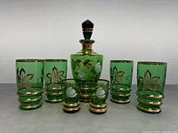 Front view of the green glass decanter with stopper and six matching glasses, showing hand painted floral and gold designs.