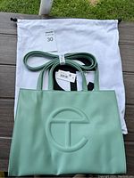 Photo of the sage green Telfar bag on a wooden surface with its white dust bag behind it, showing the front embossed logo and straps