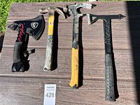 Four camping tools are laid out on a wood surface outdoors: a small axe with a sheath and three hammers with various handle colors and styles.