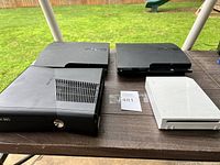 Photo showing the Xbox 360, two PS3 consoles, and Nintendo Wii on a wooden table outdoors with grass background.