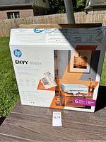 Box of HP ENVY 6055e printer in an outdoor setting showing front and side with product image and features.