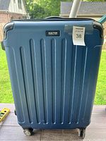 Front view of blue hard shell suitcase with vertical grooves and metal corner protectors, showing the Kenneth Cole Reaction logo and lot sticker.