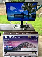 LG 43-inch 4K UHD TV displayed on a wooden table outdoors with scenic landscape on screen. TV stand and remote visible.
