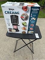 Boxed Ninja Creami frozen dessert maker on black table outdoors, with box graphics showing the product and desserts it prepares.