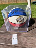 Front view of the Spalding NBA All-Star 2000 basketball in the clear acrylic case on a wooden surface outside in sunlight.
