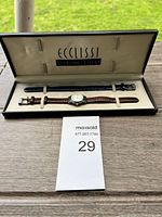 Ecclissi watch in open presentation case with two interchangeable straps (brown and blue) inside, and auction lot number card on wooden surface.