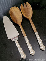 Three utensils laid out on black surface: cake server with metal blade and wooden spoon and fork with sterling silver ornate handles.