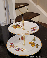 Two-tier dessert stand shown upright on a black surface with stairs in background, showing fruit motifs and gold rim.
