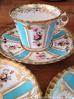 Close-up of one cup and saucer showing turquoise panels, floral bouquets, and gold scalloped rim.