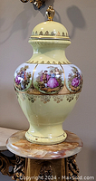 Full view of large pale yellow covered vase with painted classical figures and gold decorative accents sitting on a marble or stone pedestal.