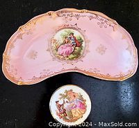 Kidney-shaped pink porcelain dish with gold trim showing seated romantic couple scene, paired with small round covered box featuring lady and dog scene