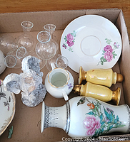 Photo of assorted porcelain saucers, clear glass decanters, yellow salt and pepper shakers, a small white jug, and a figurine.