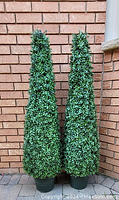 Pair of tall artificial topiary trees with dense green foliage, positioned outside against a brick wall, showing full height and detail.