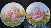 Two matching Limoges plates featuring painted scenes of a romantic couple in 18th-century clothing with nature background and gold rim detail.
