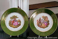 Pair of Limoges plates displayed side by side, showing the painted scenes and gold-green detailed rims.