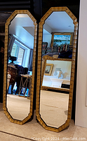 Two gilt wood framed mirrors standing against a wall, showing reflections of the room.