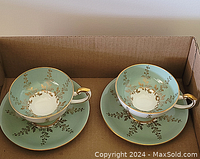 Pair of Aynsley teacups and matching saucers viewed from above inside a cardboard box, showing detailed gold leaf floral patterns on light green and white porcelain.