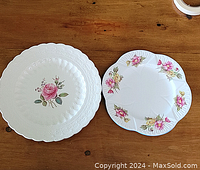 Two decorative plates on a wooden surface with floral designs, one white with embossed scalloped edges and a central rose pattern, and the other white with floral clusters around the edge.
