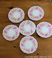 Top view of 6 white plates with the 'Serena' floral pattern featuring pink, yellow, and purple flowers with gold leaves around the rim, arranged in stacks on a wooden surface.