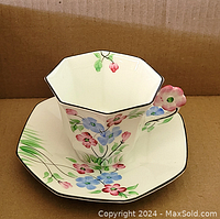 Teacup and saucer displayed with flowers painted in blue and pink, handle shaped like a pink pansy flower, edges trimmed in black