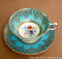 Teacup and saucer shown with turquoise blue background and gold filigree design, central floral motif on white background, ornate gold-accented handle on teacup.
