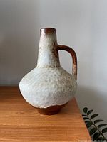 Front view of a ceramic vase showing its rounded body, narrow neck, and handle with mottled brown and beige glaze, placed on a wooden surface.